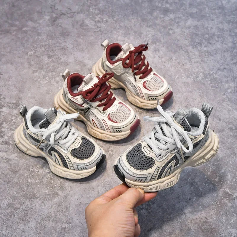 A hand holds a pair of red and white breathable mesh kids running sneakers with cushioned soles.