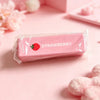 A pink strawberry shaped squishy fidget toy packaged in a clear plastic bag on a light pink surface.