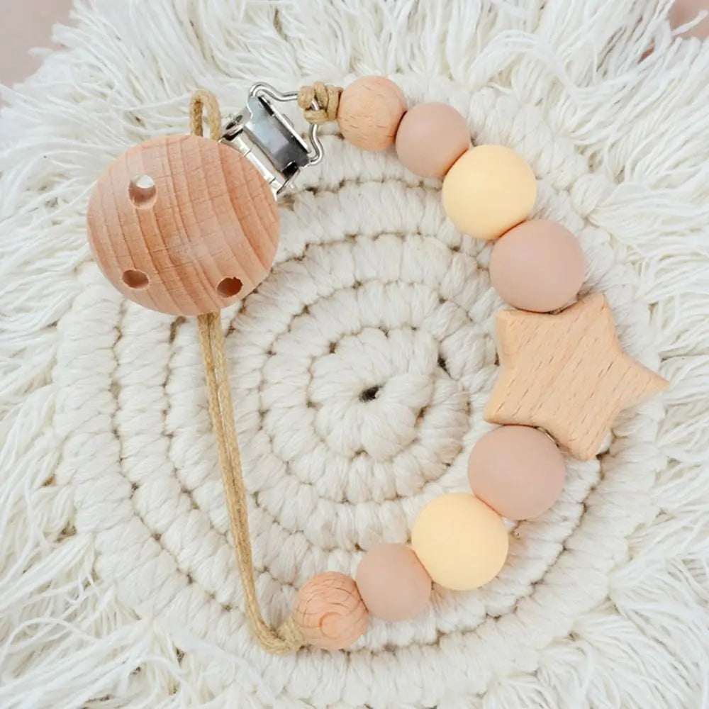 A wooden pacifier clip featuring silicone beads and a star shape, resting on a cream-colored fluffy surface.