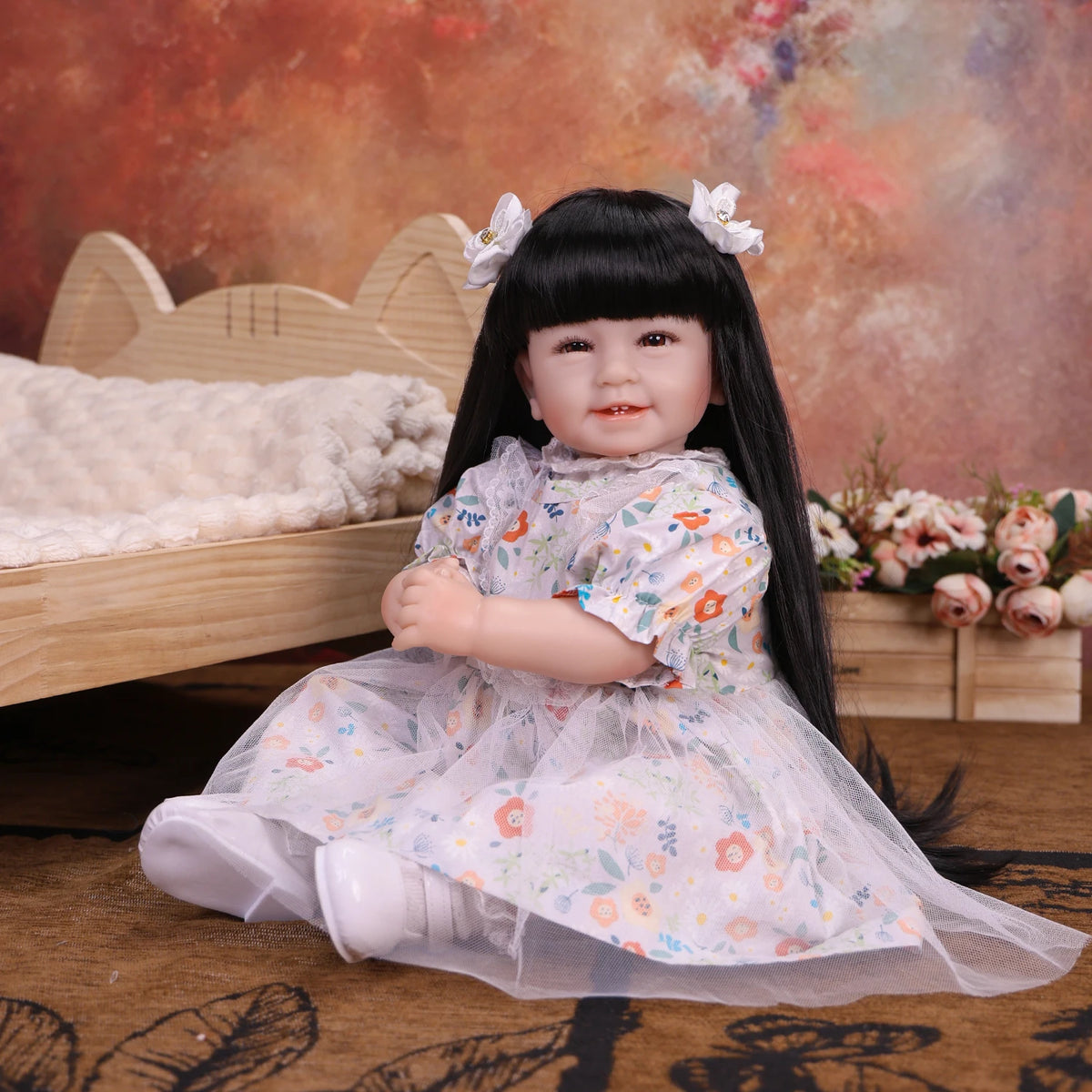 A black-haired reborn baby doll with white floral dress sitting on a wooden bed.