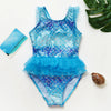 A sparkling blue and iridescent mermaid unicorn one-piece swimsuit laid on a white surface with a green leaf nearby.