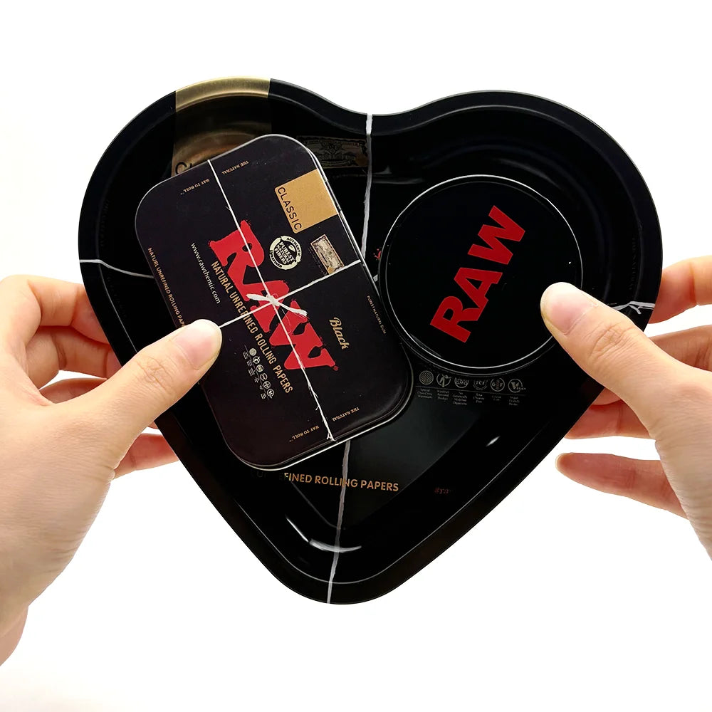 A hand holds a black love-shaped metal rolling tray with a lid open, showing a red label and a section for rolling papers.