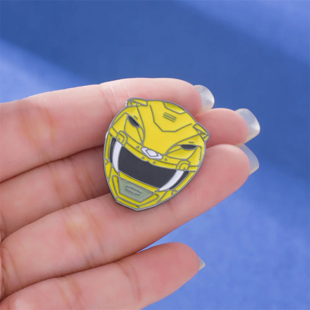 A hand holds a yellow dinosaur ranger helmet enamel pin featuring battle suit designs against a blue background.