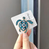 A hand holds a sparkling blue and gold rhinestone turtle brooch pin on a white card.
