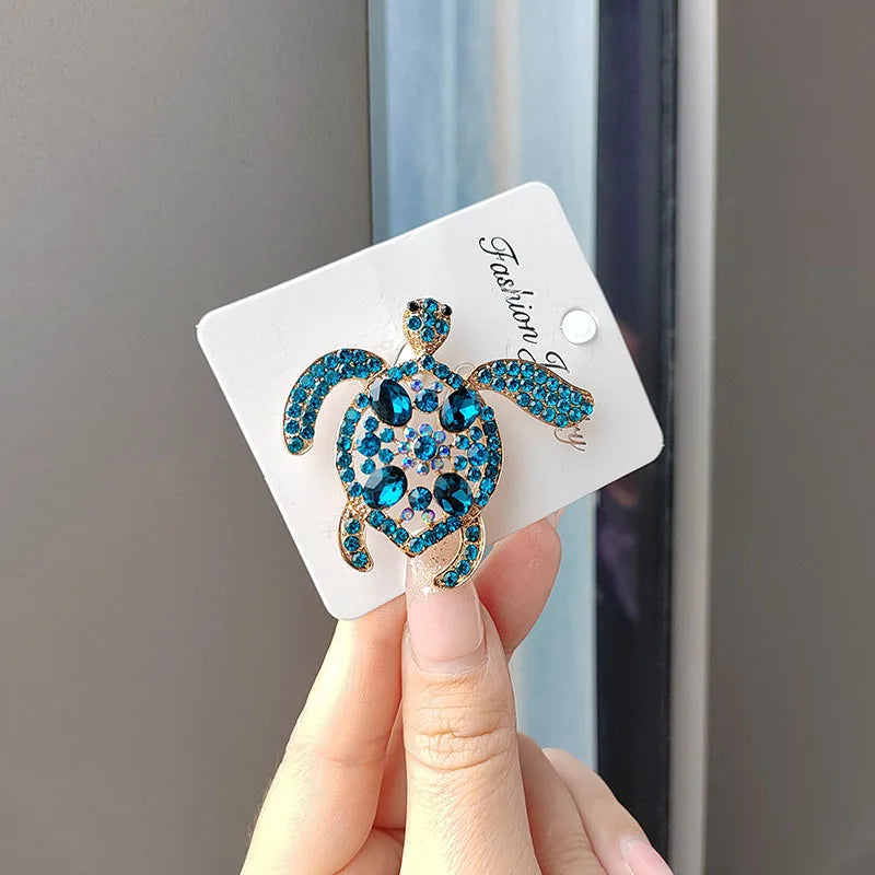 A hand holds a sparkling blue and gold rhinestone turtle brooch pin on a white card.