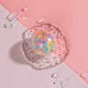 A pink and white background features a clear squishy toy filled with colorful beads, surrounded by small crystal-like pieces.