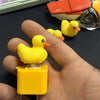 A hand holds a yellow duck fidget clicker keychain with an orange beak and LED light, next to other similar toys.