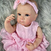 A lifelike pink reborn baby doll with painted hair wearing a ruffled dress, lying on gray fur.
