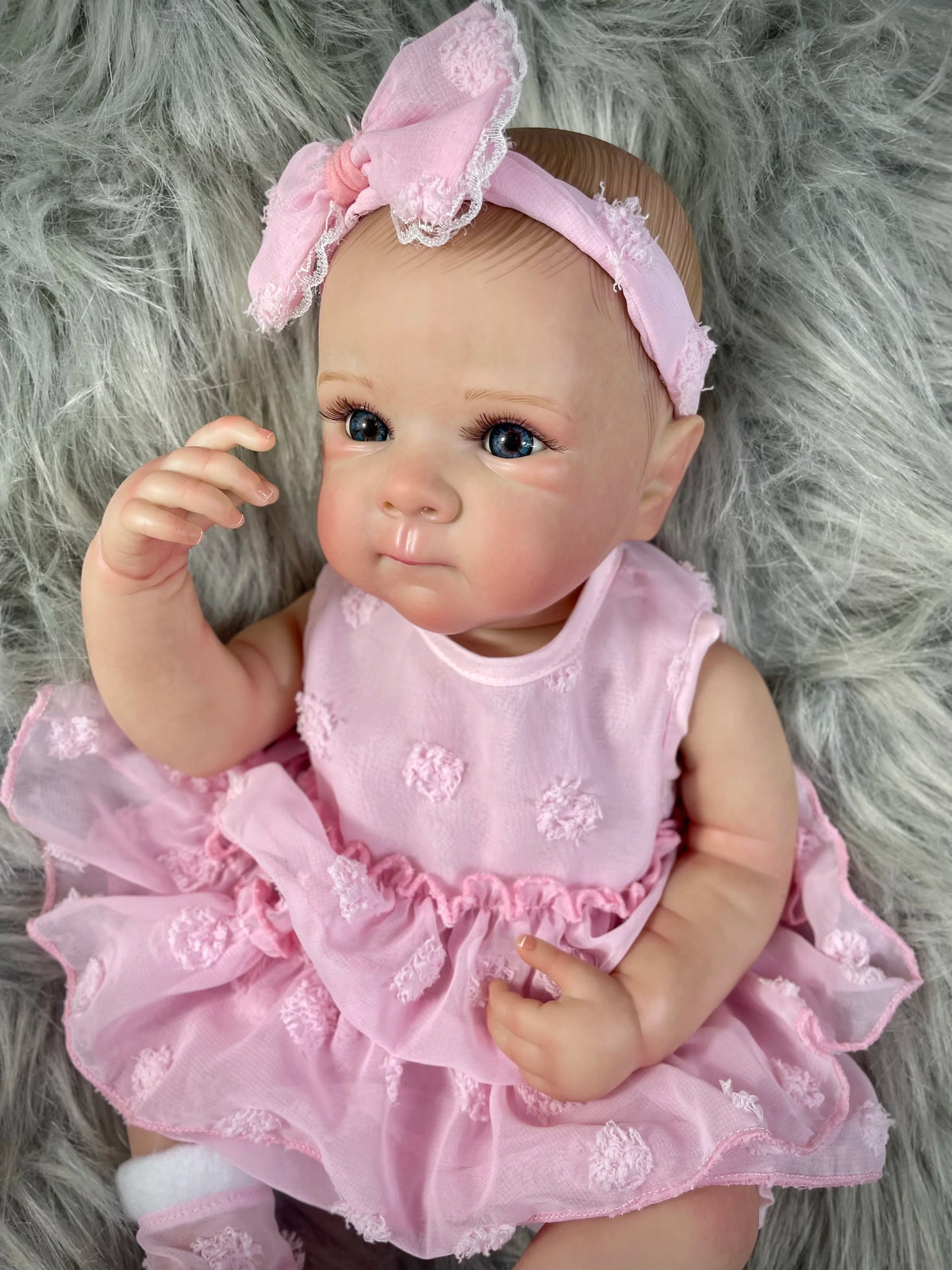 A lifelike pink reborn baby doll with painted hair wearing a ruffled dress, lying on gray fur.