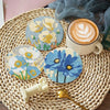 A set of floral diamond painting coasters featuring blue and yellow flowers on a woven mat with a coffee cup.