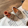 A pair of girls' brown suede slip-on moccasins with bow details resting on a white linen cloth.