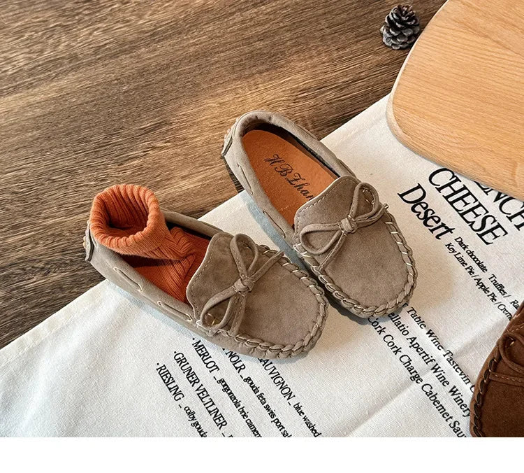 A pair of girls' brown suede slip-on moccasins with bow details resting on a white linen cloth.