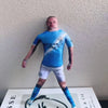 A male football star stuffed doll wearing a blue jersey and white shorts standing against a light background.