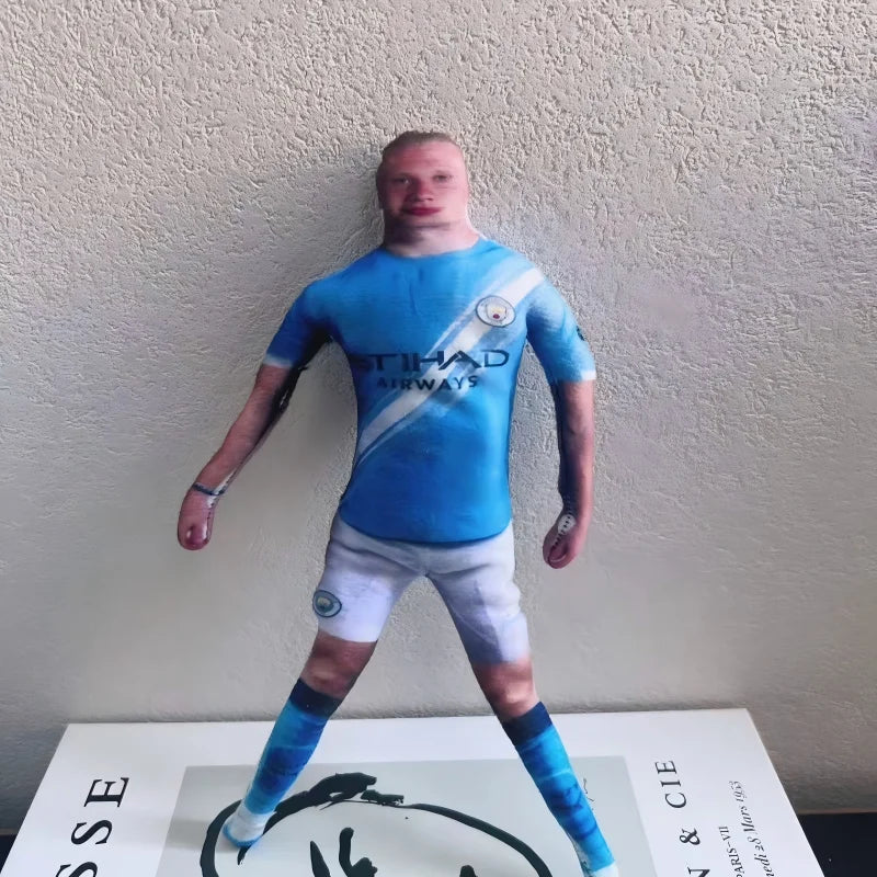 A male football star stuffed doll wearing a blue jersey and white shorts standing against a light background.