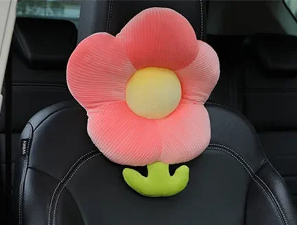 A pink plush lumbar support pillow shaped like a flower with a yellow center and green stem placed on a black car seat.