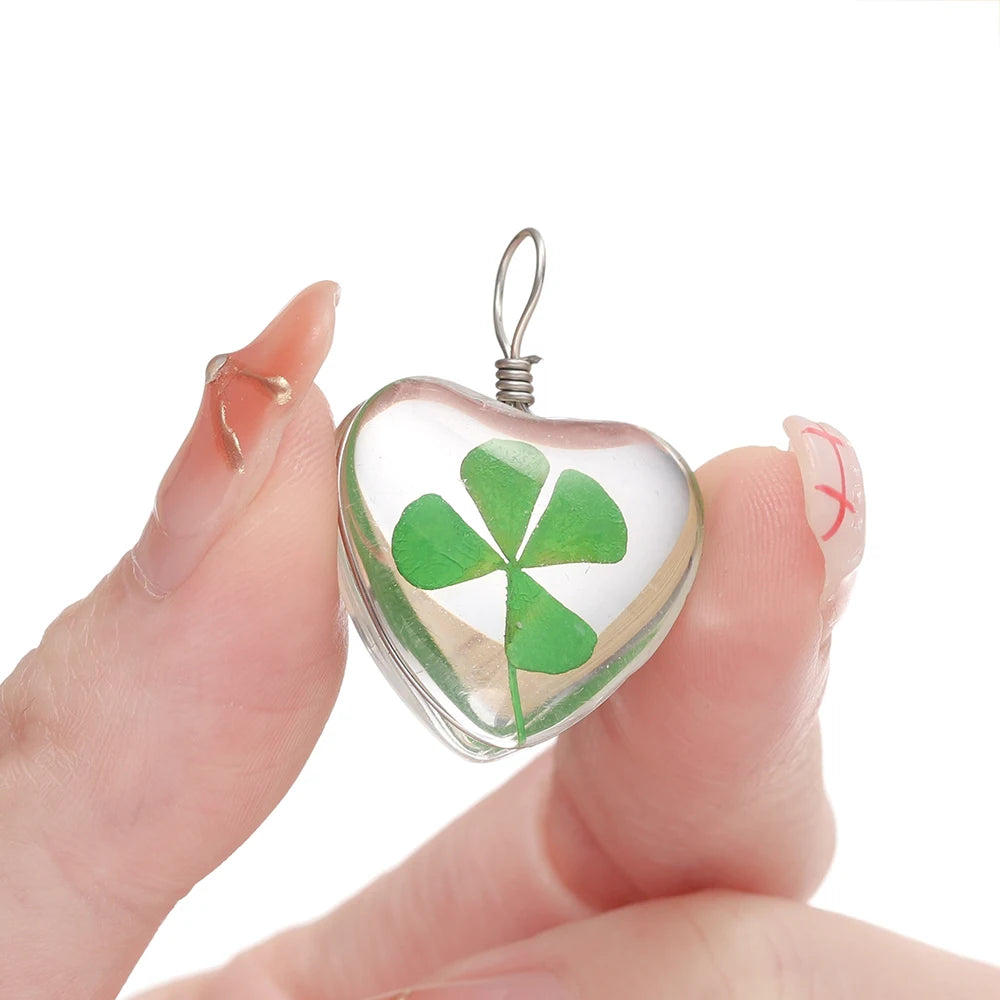 A hand holds a clear glass heart pendant with a green four leaf clover design and an attached silver loop.