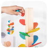 A hand is placing a blue ball onto a wooden rainbow track toy made of colorful blocks.