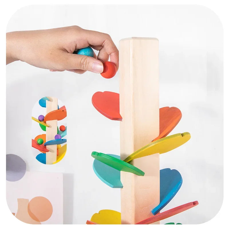 A hand is placing a blue ball onto a wooden rainbow track toy made of colorful blocks.