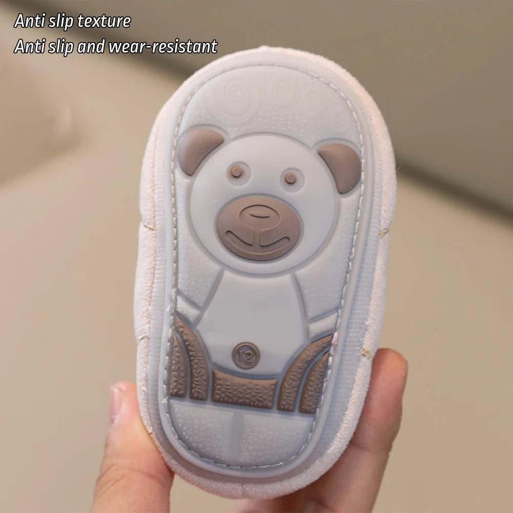 A hand holds a white baby soft sole first walker shoe featuring a bear design and anti-slip texture.