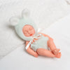 A realistic soft vinyl baby doll wearing a mint green bunny hat and outfit, lying on a white towel.