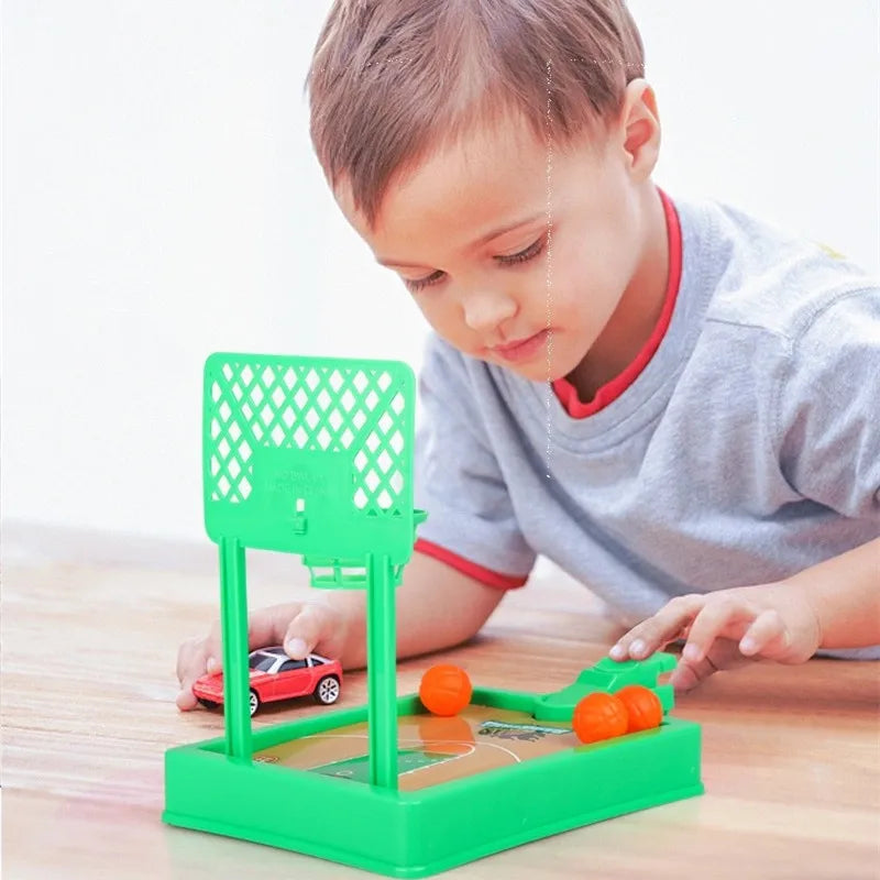 A young boy plays with a green and orange mini tabletop basketball arcade game on a wooden table.