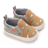 A pair of baby first walker sneakers with a tan canvas upper featuring small dinosaur prints and gray suede accents.
