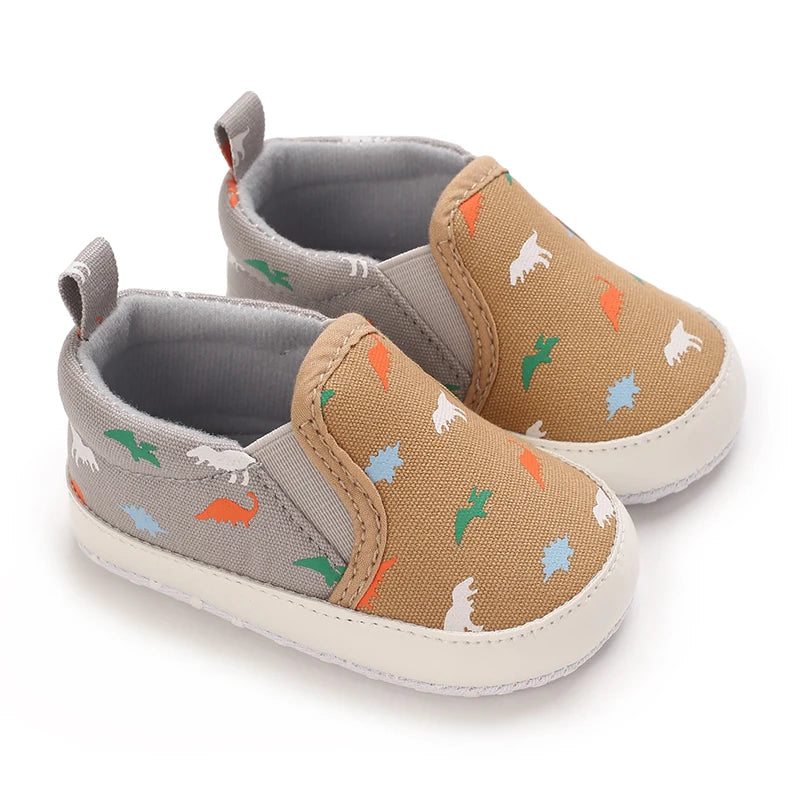 A pair of baby first walker sneakers with a tan canvas upper featuring small dinosaur prints and gray suede accents.