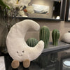 A white crescent moon plushie with a smiling face is displayed alongside various green cactus-shaped decorative items.