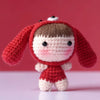 A handmade red crochet doll kit featuring a face and body, displayed against a pink background.