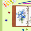 A brown watercolor sketchbook with a lay-flat binding is shown next to paintbrushes and watercolor palettes.