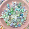 A collection of iridescent acrylic bow and flower beads in various shades of blue, green, and purple for DIY jewelry making.