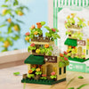 A miniature wooden building set featuring a cabin and windmill structure with flowers and greenery.