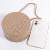 A round, beige handwoven straw crossbody bag with a natural fiber strap and lace bow next to a white smartphone.