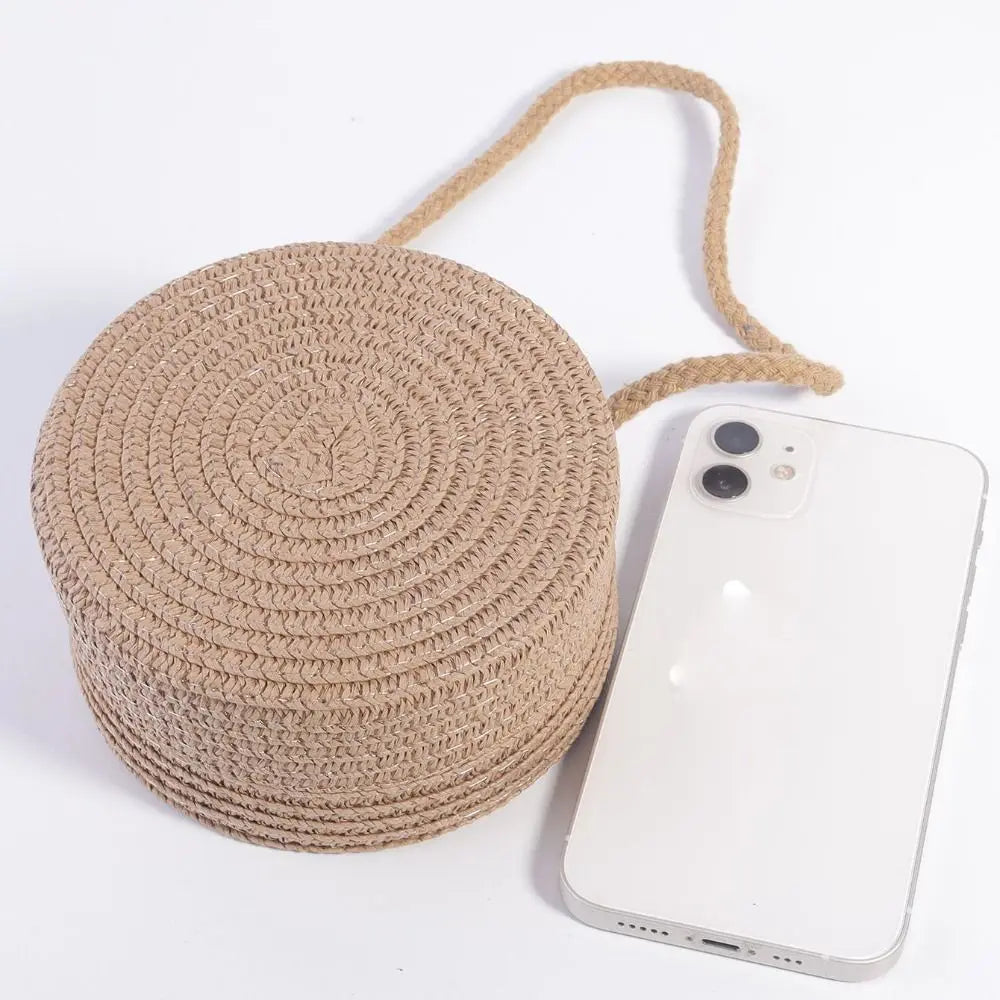 A round, beige handwoven straw crossbody bag with a natural fiber strap and lace bow next to a white smartphone.