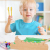 A toddler holds a wooden test tube clip beads sorting toy in front of a white table.