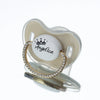 A clear silicone baby pacifier with a light beige crown-shaped mold and a gold rhinestone ring displaying the name Angelica.