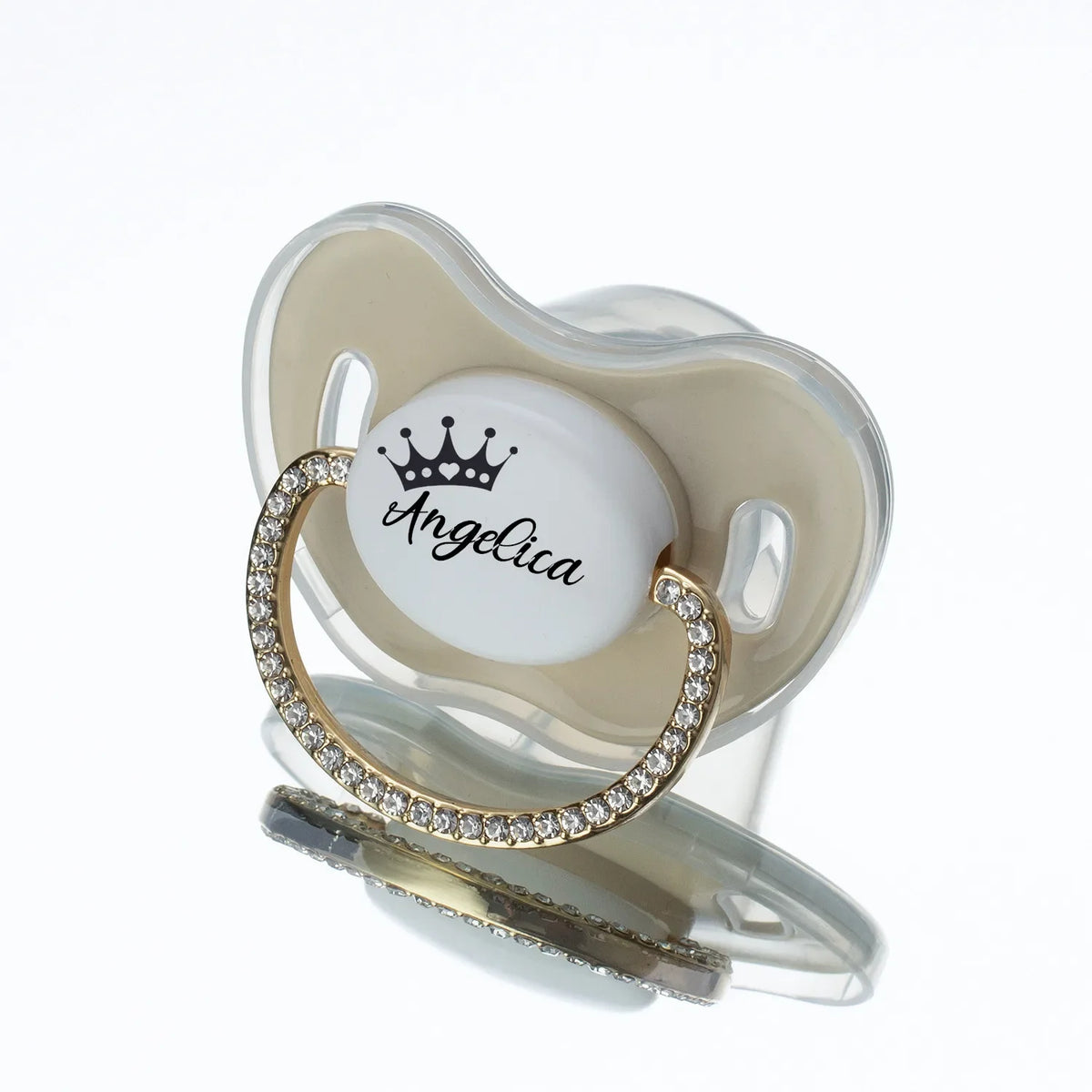 A clear silicone baby pacifier with a light beige crown-shaped mold and a gold rhinestone ring displaying the name Angelica.