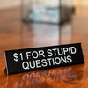 A black desk sign with white text that reads $1 for stupid questions placed on a wooden surface.