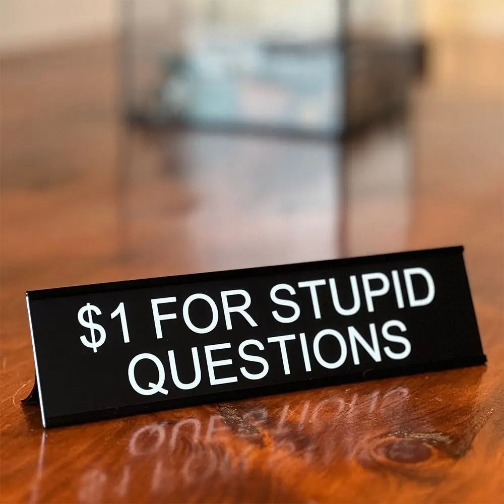 A black desk sign with white text that reads $1 for stupid questions placed on a wooden surface.