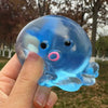 A hand holds a translucent blue stretchable octopus stress ball with a pink ring on a park background.
