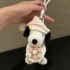 A white Snoopy plush charm with a pink hat and a floral wreath is being held by a hand.