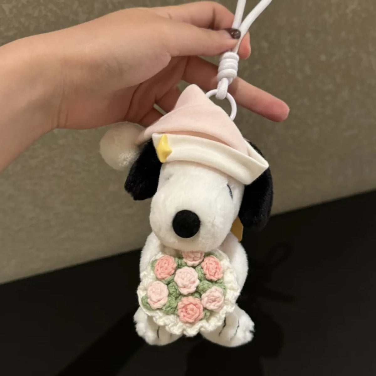 A white Snoopy plush charm with a pink hat and a floral wreath is being held by a hand.