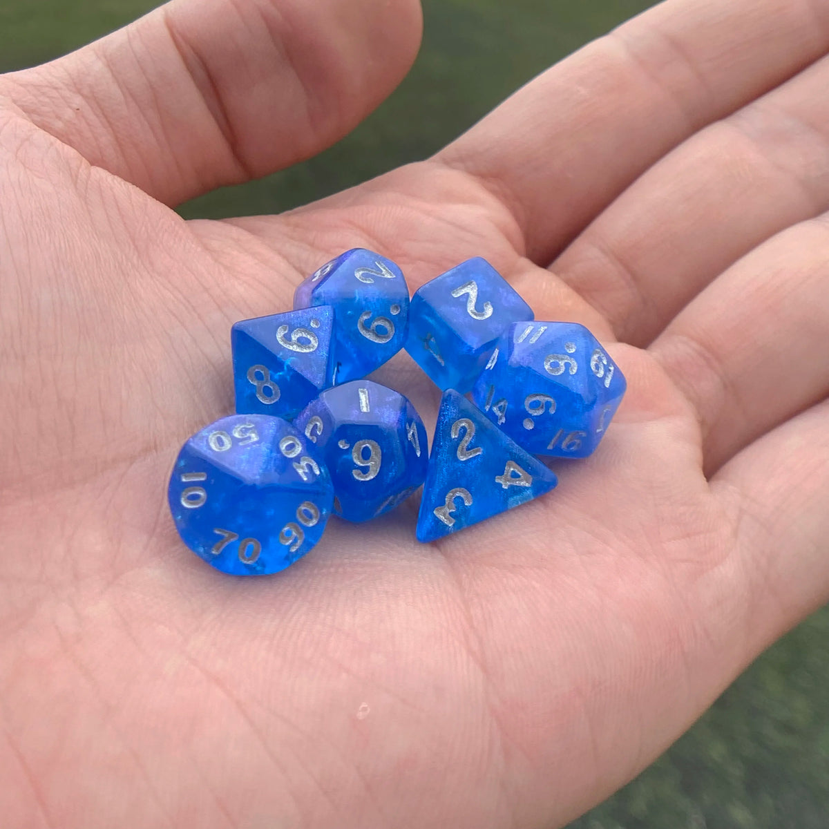 A hand holds a set of seven iridescent blue acrylic polyhedral dice on a green background.