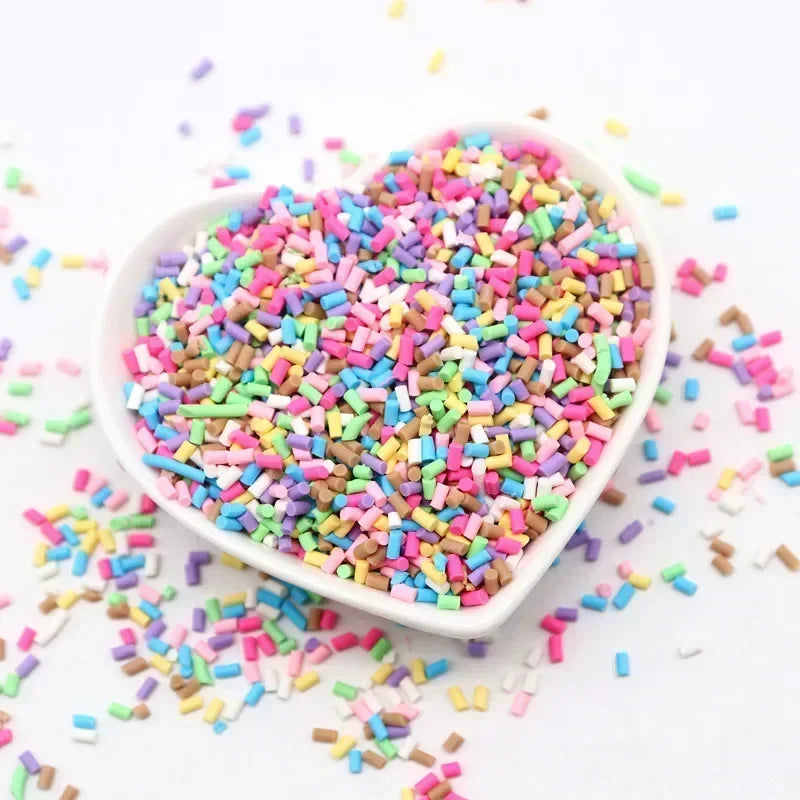 A heart-shaped dish filled with colorful polymer clay sprinkles for slime and craft decor scattered on a white surface.
