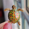 A small turtle brooch pin with iridescent green and yellow scales and gold accents is held by a hand.