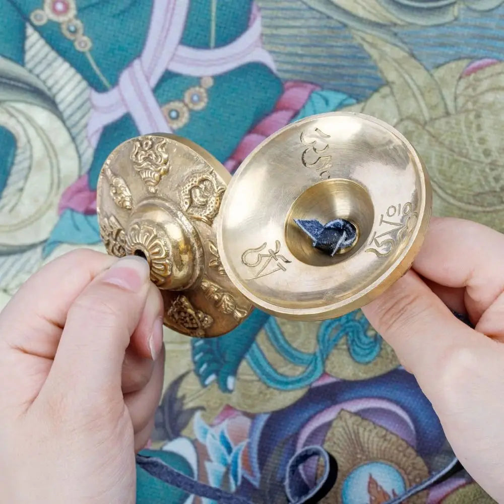 A hand holds a handcrafted brass Tingsha cymbal featuring eight auspicious symbols against a patterned fabric.