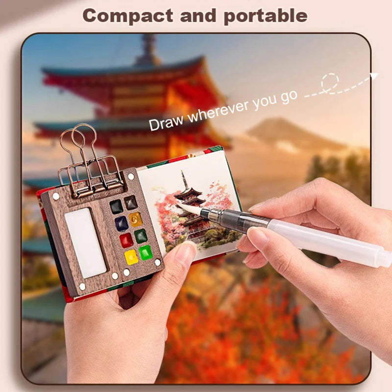 A hand draws on a portable wooden watercolor paint set outdoors against a scenic background.