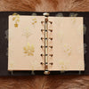 A page from a notebook featuring delicate gold botanical illustrations on light beige paper, bound with a brass spiral bindin