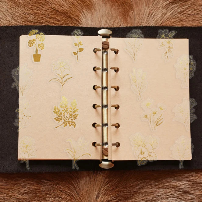 A page from a notebook featuring delicate gold botanical illustrations on light beige paper, bound with a brass spiral bindin