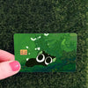 A green card with a cute black cat design is held against a dark green grassy background.